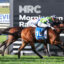 Two jockeys ride racehorses at full speed, necks stretched toward the finish line, with a prominent MRC sign behind them.