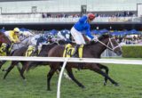 Jockey in blue silks on a dark brown horse leading a horse race on a grassy track with cheering spectators in the background.