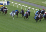 Group of jockeys riding horses in a crowded horse race on a grassy track with white rails. colorful silks contrast against green turf, timer on screen.