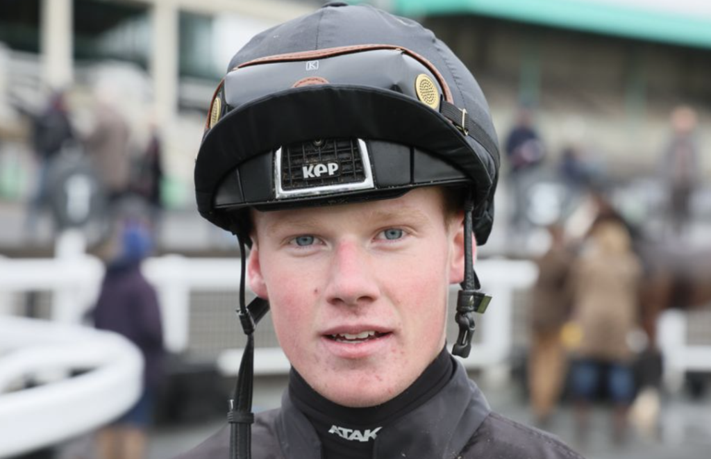 Close-up portrait of a young jockey in a black helmet and dark racing silks, looking at the camera with a blurred crowd in the background.