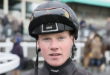 Close-up portrait of a young jockey in a black helmet and dark racing silks, looking at the camera with a blurred crowd in the background.