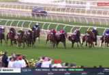 Horses sprinting on a green turf racecourse with colorful jockey silks, crowd watching from the infield rail as they race toward the finish.