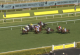 Group of racing horses with jockeys in colorful silks competing along the inside rail on a green turf track.
