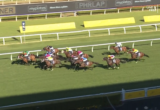 Horses and jockeys sprinting on a green grass racetrack during a daytime horse race.
