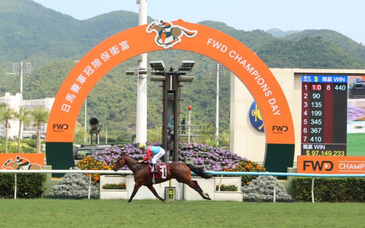 Horse and jockey racing past the finish line under an orange FWD champions arch with a digital scoreboard nearby and mountains in the background.