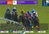 Group of Thoroughbred horses and jockeys racing on a green turf track, bunching together mid-turn with sponsor banners overhead and on-screen race data around the edges.