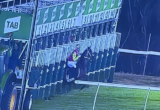 Jockey in bright pink and yellow silks stands beside the starting gate at a horse race, with numbered stalls and a TAB sign in the background.