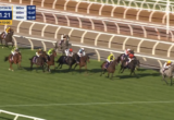 Horse race on a turf track with multiple jockeys in colorful silks, horses galloping near the inside rail; on-screen timer reads 2:41.21 and '220m to go' with race graphics in the corner.