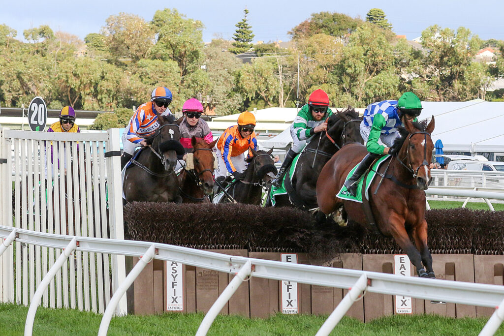Jockeys and horses jump over a hurdle in a horse race, colorful silks visible against a backdrop of trees and blue sky.
