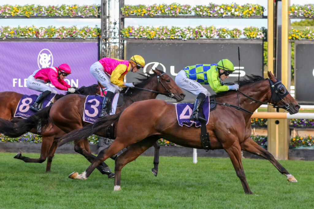 Three jockeys in bright pink, yellow, and green silks race side by side on a grassy turf track, leaning forward in full sprint.