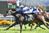 Group of racehorses and jockeys sprint toward the finish line on a sunny track, blue and white silks leading, scoreboard in the background showing time.