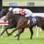 Jockeys lean forward riding racehorses as they sprint on a grassy track at a close finish behind a red rail fence.