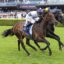 Two jockeys ride fast on a grassy racecourse, the brown horse in white-and-blue silks leading ahead of a dark horse with a crowd and grandstands behind them.