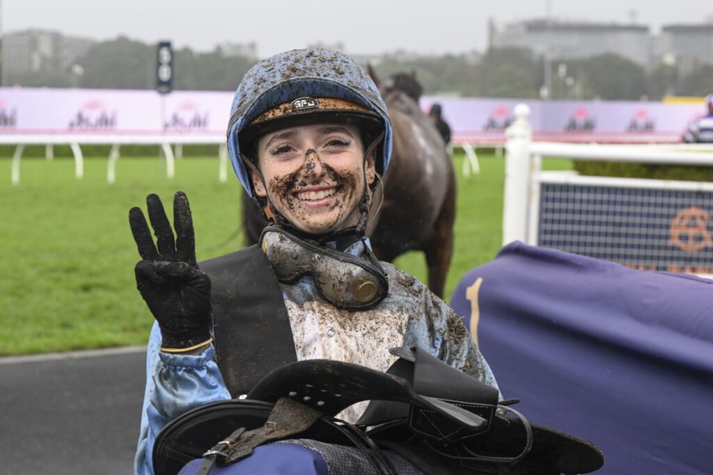 Siena Grima | Just Horse Racing Jockey in blue silks and helmet, muddy face and gear, flashing a peace sign after a race on a track in the rain.