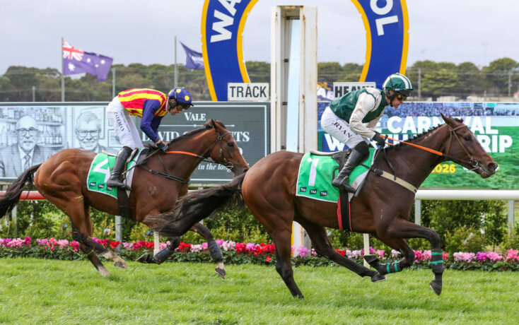 Two jockeys race side by side on galloping horses on a green turf track, with a large blue-and-yellow arch and advertising boards in the background.