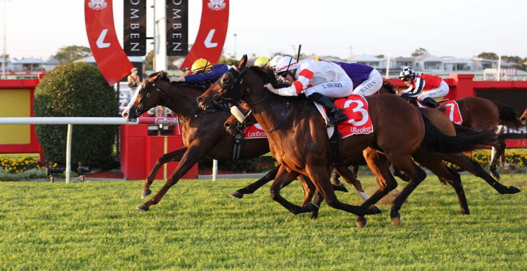 Horses and jockeys sprinting side by side in a crowded horse race on a green track with red fences and banners in the background