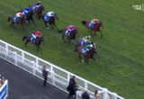 Horses racing on a green turf course, jockeys in bright colorful silks in a pack near the railing, viewed from above at the trackside slope.
