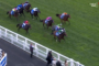 Horses racing on a green turf course, jockeys in bright colorful silks in a pack near the railing, viewed from above at the trackside slope.
