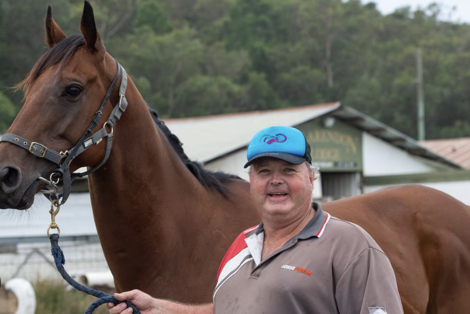 Brett Bellamy | Just Horse Racing Man in a gray polo holding a lead attached to a brown horse at a stable outdoors.