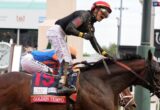 Jockey in red helmet riding a dark horse mid-jump in a horse race, with a blue-clad rider behind; saddle cloth reads GOLDEN TEMPO.
