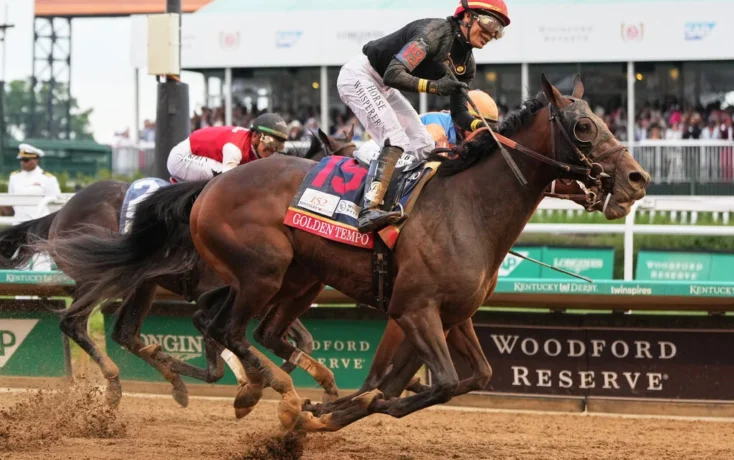 A straining jockey in a red helmet rides a dark brown racehorse at full speed on a dirt track, ahead of rivals at Woodford Reserve Downs, as spectators watch in the background.