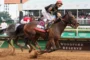 A straining jockey in a red helmet rides a dark brown racehorse at full speed on a dirt track, ahead of rivals at Woodford Reserve Downs, as spectators watch in the background.