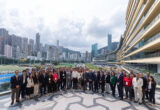 Group of professionals in business attire posing for a photo on a rooftop terrace with a city skyline and a curved modern building on the right.