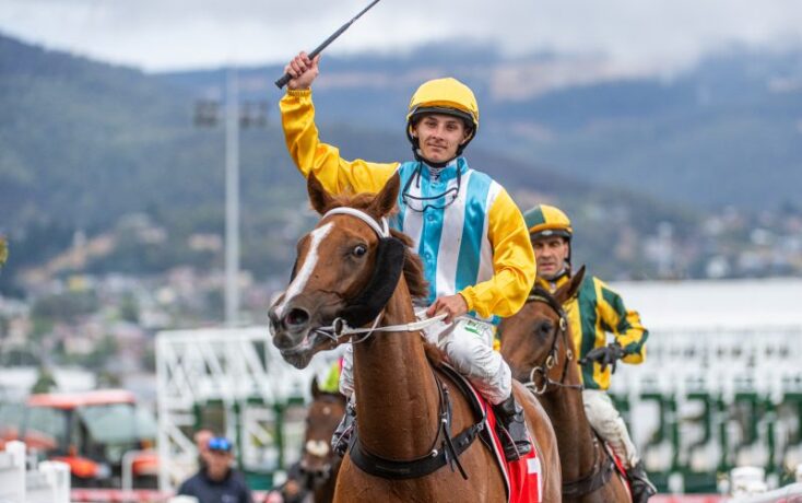 Jockey in a yellow helmet and blue-striped vest raises a whip while riding a brown horse; another rider in green and yellow follows behind at a race track with hills in the background.