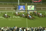 Horses and jockeys race toward the finish line at Aquis Park Gold Coast, with a cheering crowd in the foreground.