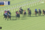 Jockeys in colorful silks race side-by-side on a green turf track near the inside rail, moment of a tight finish near the front of the group.