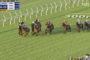 Horses with colorful jockeys race on a green turf track near the 300m mark beside a white railing.