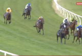 Horses race on a grass track with jockeys in colorful silks leaning forward in the lead group near the rail.