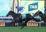 Two jockeys ride galloping horses in a race, with the lead horse wearing a green saddlecloth and the rider in blue diamond-pattern silks ahead of a rival.