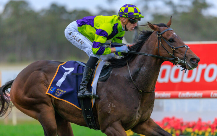 Jockey wearing neon green and purple silks rides a brown horse during a race, leaning forward with goggles on.