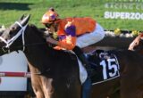 Jockey in orange and purple silks rides a racing horse with number 15 on a blue saddle cloth during a race track event.