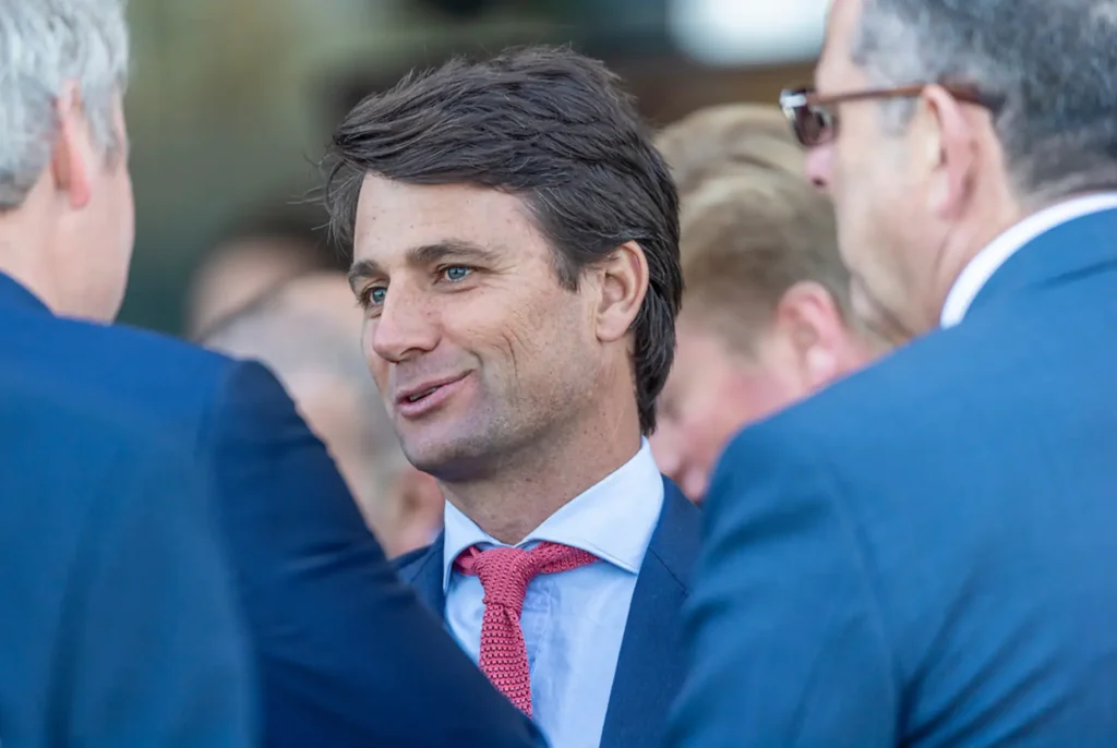 Man in a navy suit and red tie smiles while conversing with others at a formal event.