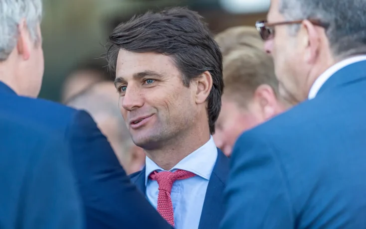 Man in a navy suit and red tie smiles while conversing with others at a formal event.