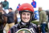 Smiling female jockey in a red helmet holds a silver trophy, with blurred crowd and horses in the background.