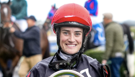 Smiling female jockey in a red helmet holds a silver trophy, with blurred crowd and horses in the background.