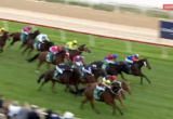 Horse race: jockeys in colorful silks ride thoroughbreds on a green turf track, spectators blur on the left, Racing.com live logo in the corner.