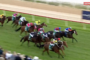 Horse race: jockeys in colorful silks ride thoroughbreds on a green turf track, spectators blur on the left, Racing.com live logo in the corner.