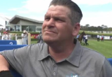 Man in a gray polo leaning on a blue railing at a horse racetrack, with spectators and horses visible in the background on a sunny day.