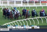 Horse race on a green turf track with a large field of jockeys in colorful silks rounding a bend, rails visible.