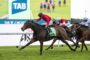 Horse race in progress: leading brown horse with green saddlecloth is ahead of two trailing horses on a grassy track under a large TAB banner screen.