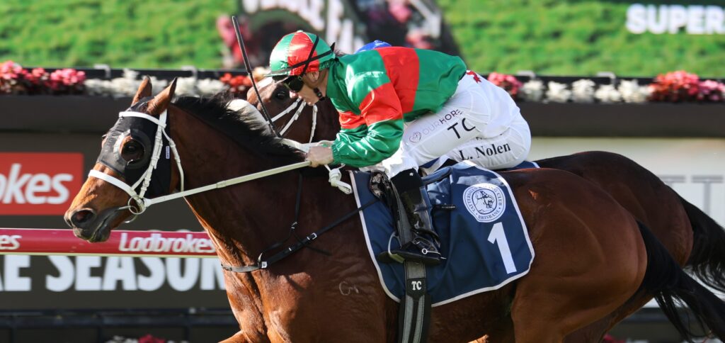 Jockey in green and red silks leans forward on a brown racehorse during a race.