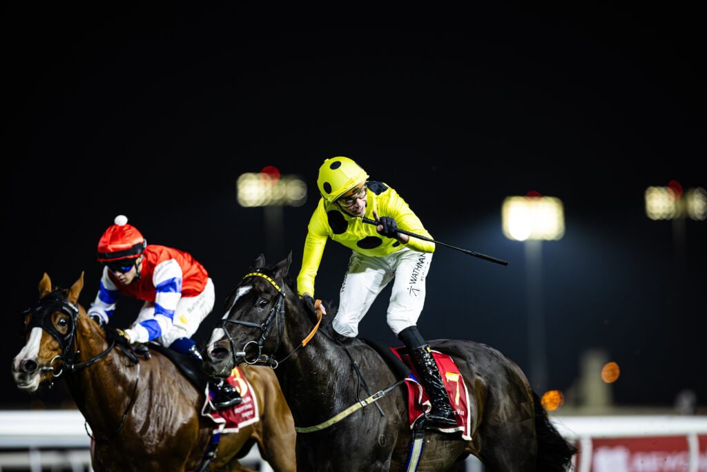 Jockey in bright yellow polka-dot silks riding a dark horse at a nighttime horse race, other riders nearby.