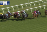 Group of horse racehorses galloping on a grass track with jockeys in colorful silks behind white railing. A close pack leads toward the bend under a sunny sky, timer and banners visible on screen edge.