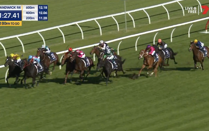 Group of horse racehorses galloping on a grass track with jockeys in colorful silks behind white railing. A close pack leads toward the bend under a sunny sky, timer and banners visible on screen edge.