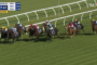 Group of horse racehorses galloping on a grass track with jockeys in colorful silks behind white railing. A close pack leads toward the bend under a sunny sky, timer and banners visible on screen edge.