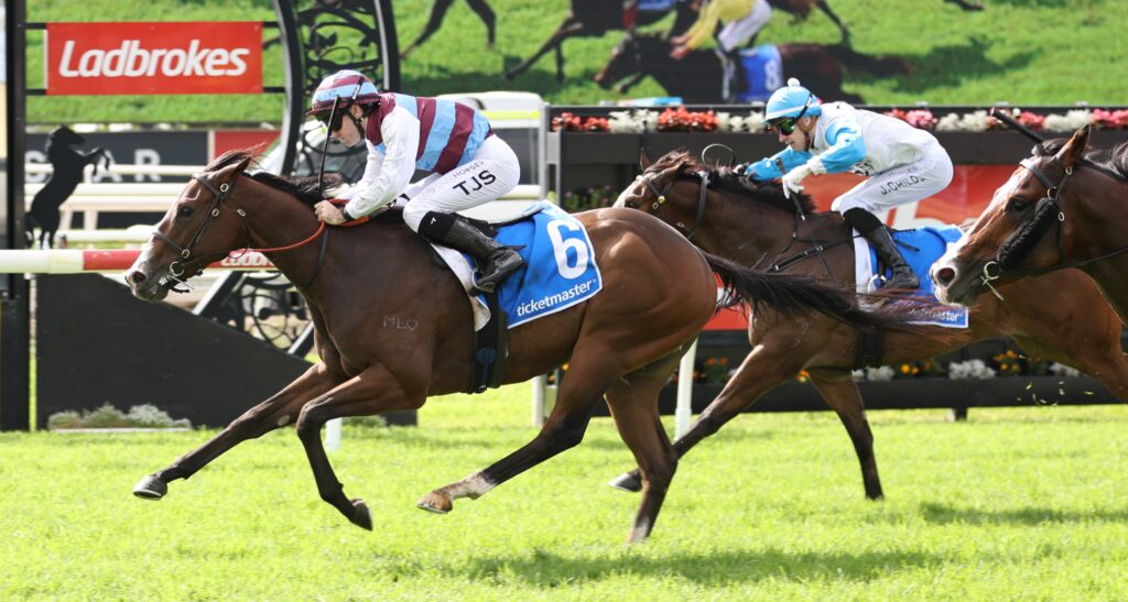 Two jockeys ride racehorses at a track, with a blue saddle cloth labeled 6 in the foreground and a Ladbrokes banner in back.